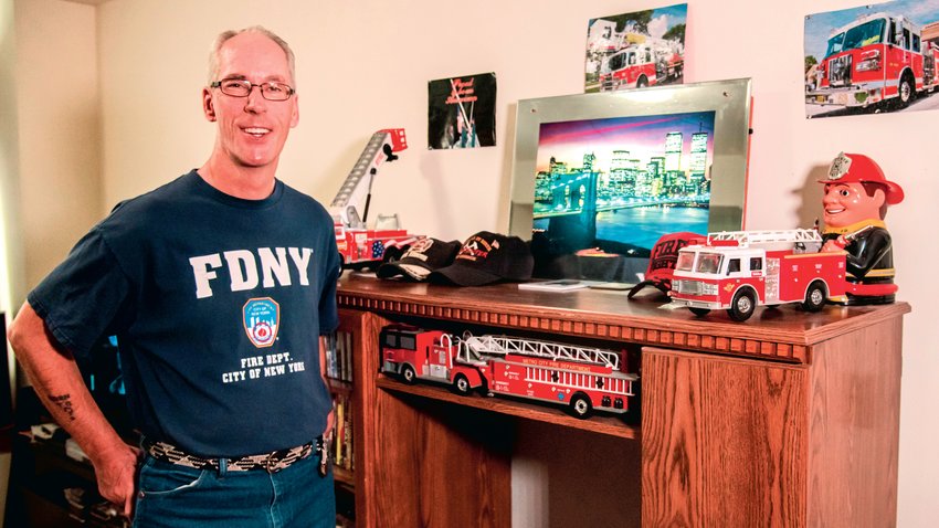 Charles Tippett Jr. smiles and poses for a photo next to a collection of fire hats and engines displayed next to an illuminated photo of the New York City skyline in his Centralia residence. Tippett Jr. sports an &ldquo;FDNY&rdquo; shirt he received while working as a firefighter in the city, where he helped victims during the events that unfolded on Sept. 11, 2001. &quot;That's my boys right there,&quot; he said Saturday while describing the fire trucks displayed in his living room.