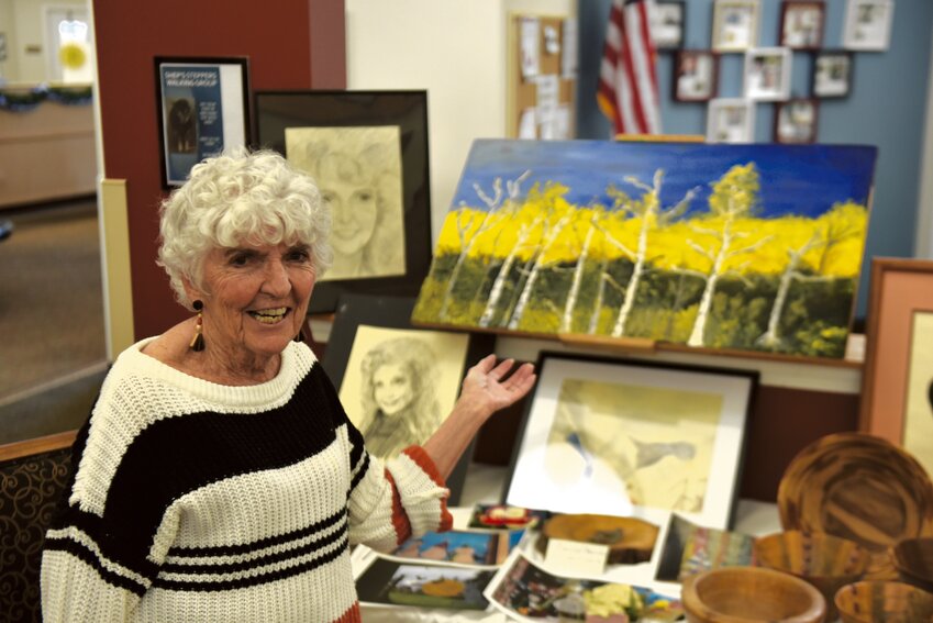 Prestige Senior Living Rosemont resident Carolyn Benum smiles and poses next to her artwork at the facility's art walk on May 10.
