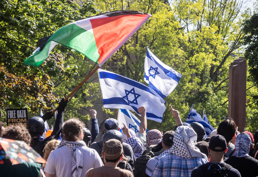 Seen from behind the first line inside the pro-Palestinian encampment on the Quad at the University of Washington, fists go up under a Palestinian flag, Sunday, May 12, 2024 in Seattle, as the group shouts against counter-demonstrators waving Isreali flags, just across the barricade.