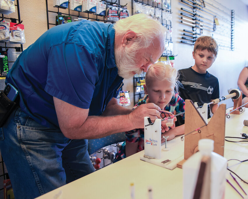 Richard Bowers, left, shows Ellianna Fraidenburg, middle, how to apply epoxy to the pink threading on her fishing rod as her brother, Elias, watches closely during a class teaching youth how to build their own fishing rods at Battle Ground Rod n Reel on Wednesday, July 24.